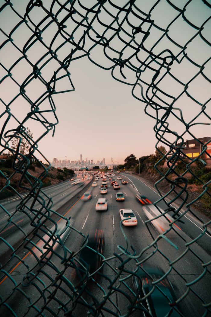 Traffic flows on a highway framed by a broken fence with San Francisco cityscape at sunset.